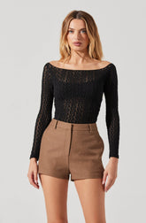 A woman is wearing a black, long-sleeved, open-knit top with a wide neckline that exposes her shoulders, paired with high-waisted, tailored brown shorts.